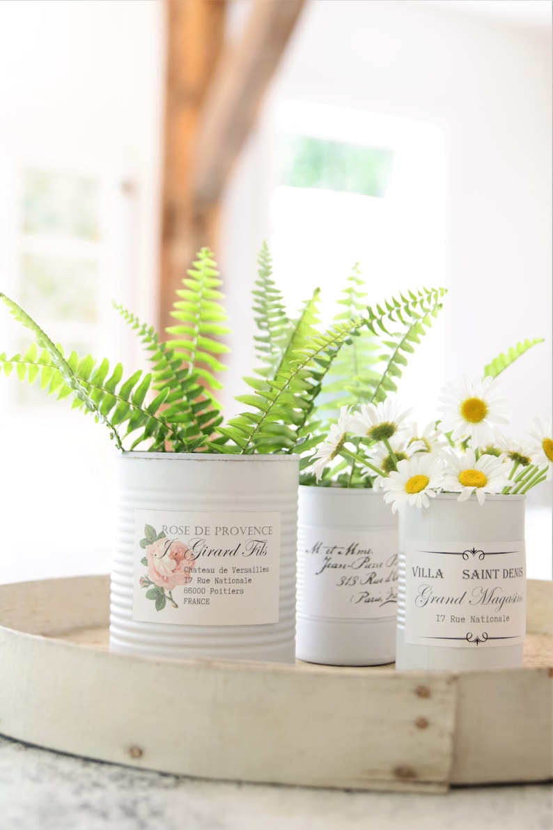 May include: Three white metal cans with labels and decorative designs. The cans are filled with green ferns and white daisies. The cans are sitting on a white tray.