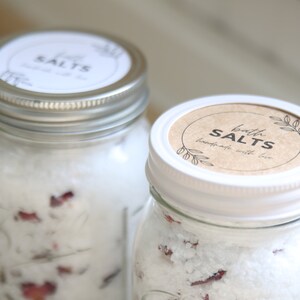 May include: Two glass jars filled with white bath salts and dried rose petals. The jars have white lids with brown labels that read "bath salts handmade with love".