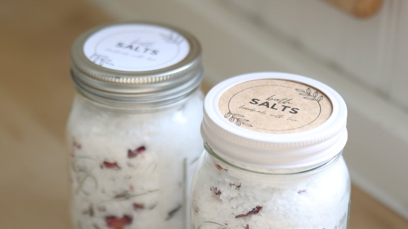 May include: Two glass jars filled with white bath salts and dried rose petals. The jars have white lids with brown labels that read "bath salts handmade with love".