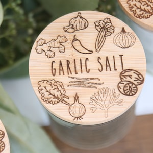 K&ouml;nnte beinhalten: Nahaufnahme eines Bambusdeckels mit dem Schriftzug "GARLIC SALT" in der Mitte. Der Deckel ist mit ge&auml;tzten Illustrationen von Knoblauch, Ingwer, Chili, Sellerie, Zwiebel, Muskatnuss und anderen Kr&auml;utern verziert. Der Deckel befindet sich auf einem wei&szlig;en Glas.