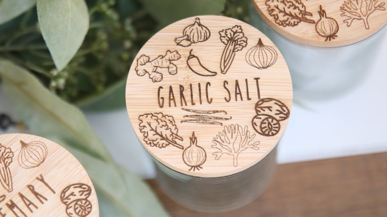 K&ouml;nnte beinhalten: Nahaufnahme eines Bambusdeckels mit dem Schriftzug "GARLIC SALT" in der Mitte. Der Deckel ist mit ge&auml;tzten Illustrationen von Knoblauch, Ingwer, Chili, Sellerie, Zwiebel, Muskatnuss und anderen Kr&auml;utern verziert. Der Deckel befindet sich auf einem wei&szlig;en Glas.