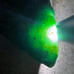 May include: A raw, translucent green mineral specimen with an irregular, jagged shape. A bright white light source is directed at one side, illuminating the interior and revealing deep emerald tones and textured, crystalline surfaces.
