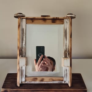 May include: A rectangular mirror in a rustic wooden frame. The frame has a distressed, two-tone finish, with natural wood and white painted accents. The mirror sits on a dark stained wooden base, creating a decorative accent piece.