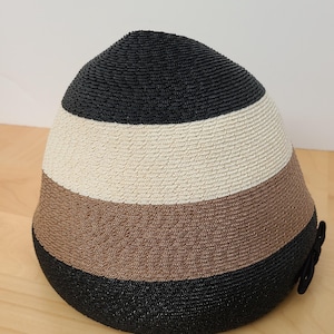 May include: A striped hat with a rounded crown. The hat features horizontal bands of black, off-white, and brown. A small black bow is attached to the side. The hat is made of woven material and sits on a wooden surface.
