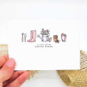 May include: A white card with a note that says "a note from LOUISA EVANS". The card has a hand-drawn illustration of gardening tools, pink rain boots, a watering can, flowers, and two small pots.