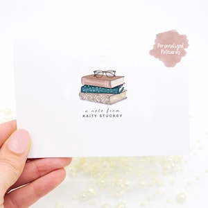 May include: A white notecard with a stack of three books, a pair of glasses, and the text "a note from KAITY STUCKEY". The notecard has a pink watercolor background with the text "Personalized Notecards" in the upper right corner.