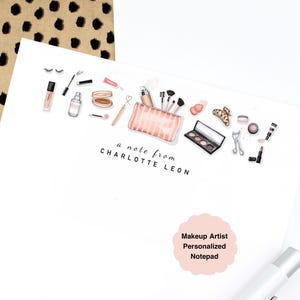 May include: A white notepad featuring illustrations of makeup products, including brushes, lipsticks, and eyeshadow. The text "a note from CHARLOTTE LEON" is printed below the illustrations. A pink sticker reads "Makeup Artist Personalized Notepad".