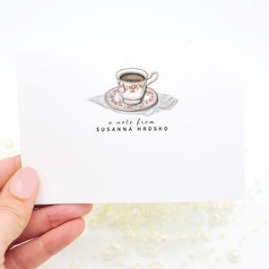 May include: A white teacup with pink and yellow floral pattern, filled with brown liquid, sits on a matching saucer with a white doily. The text "a note from SUSANNA HROSKO" is printed below the teacup.