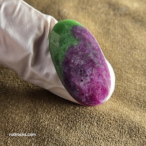 May include: An oval-shaped gemstone with a vibrant color split. The top half is a deep green, transitioning to a rich, purplish-red hue on the bottom. The polished stone is held by a gloved hand, showcasing its natural beauty.