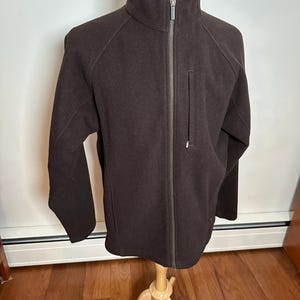 May include: A brown fleece jacket with a zipper closure and a small pocket on the left side. The jacket has a stand-up collar and long sleeves.