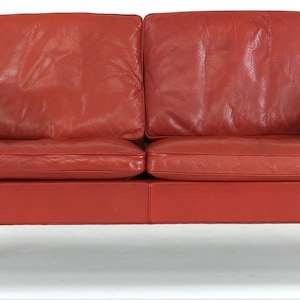 May include: A two-seater red leather sofa with a mid-century modern design. The sofa has a rectangular shape with a low profile, wooden legs, and a tufted seat and back. The overall dimensions are approximately 60 inches wide.