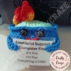 Puede incluir: Un contenedor de basura azul tejido a mano con ojos de botón negros y una llama roja y amarilla en la parte superior. El contenedor contiene una tarjeta que dice "Emotional Support Dumpster Fire. It's fine, I'm fine, Everything is FINE!"