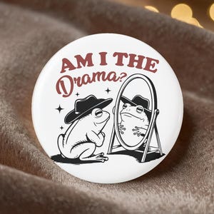 May include: A white button with a black and white illustration of a frog wearing a hat, looking in a mirror. The text "AM I THE Drama?" is in maroon and black.