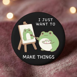 May include: A black round button featuring a cartoon frog painting a picture of itself on a small easel. The frog holds a paintbrush and a palette. The text "I JUST WANT TO MAKE THINGS" is written on the button.