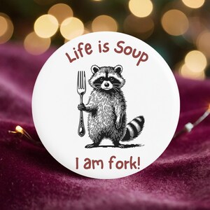 May include: A white button with a raccoon illustration holding a fork. The text "Life is Soup, I am fork!" is printed around the raccoon. The button is set against a dark red velvet background with bokeh lights.