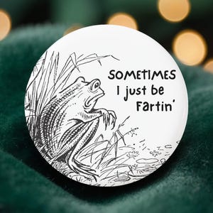 May include: A white button featuring a black and white illustration of a frog sitting in tall grass. The text on the button reads "SOMETIMES I just be Fartin'". The button is round and appears to be made of metal.