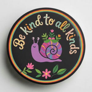 May include: A round black pinback button featuring a purple snail with a colorful striped shell. The snail carries green plants on its back, surrounded by pink flowers. The text "Be kind to all kinds" arches above in a rainbow-colored font.