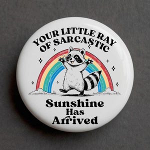 May include: A round white pin-back button featuring a black and white illustration of a raccoon standing in front of a colorful rainbow. The text reads: YOUR LITTLE RAY OF SARCASTIC Sunshine Has Arrived, surrounded by small black star icons.