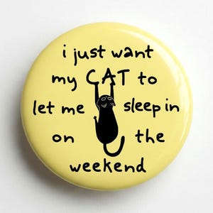 May include: A round, pale yellow pin-back button featuring a black silhouette of a cat hanging from the word CAT. The text reads: i just want my CAT to let me sleep in on the weekend, written in a simple, dark, handwritten-style font.