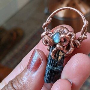 May include: A copper wire octopus with a blue gemstone in the center, holding a black tourmaline crystal pendant.