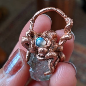 May include: A clear quartz crystal wrapped in copper wire with a blue labradorite gemstone accent. The crystal is held in a copper wire cage.