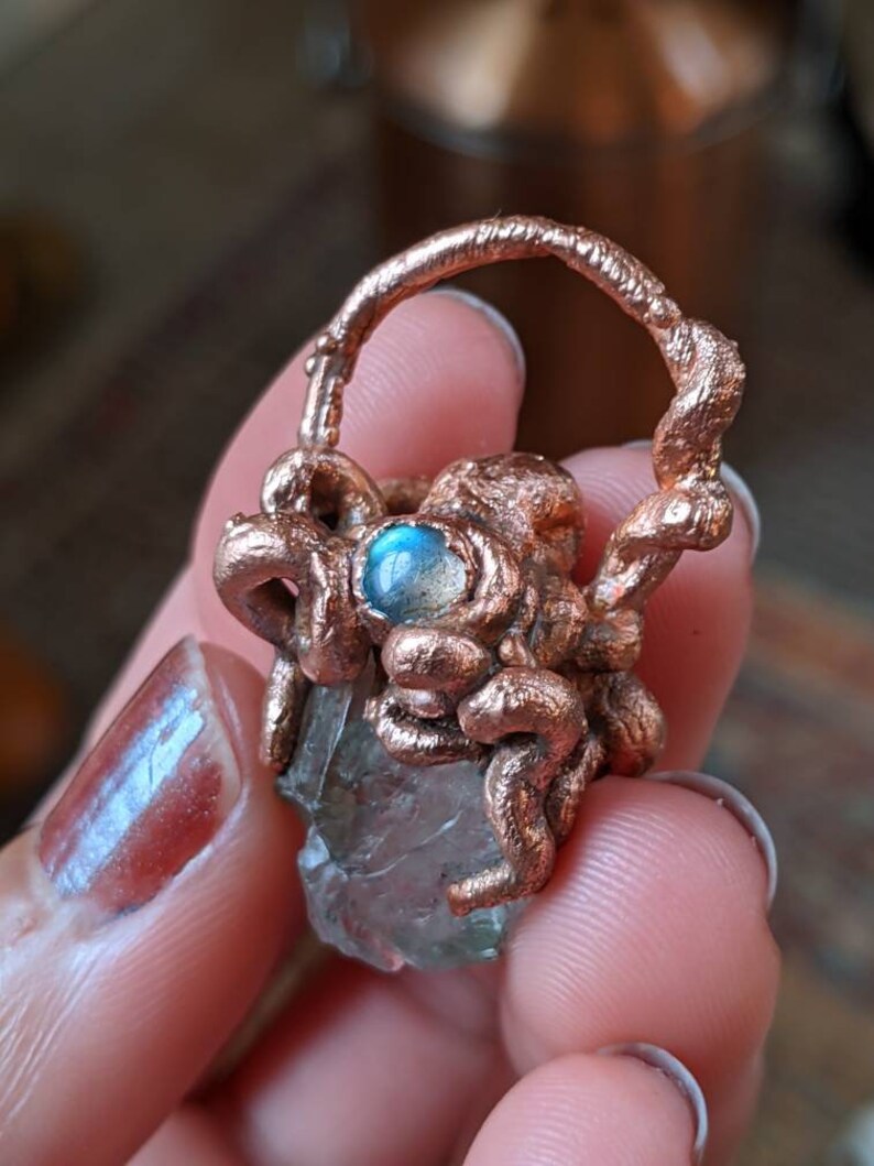 May include: A clear quartz crystal wrapped in copper wire with a blue labradorite gemstone accent. The crystal is held in a copper wire cage.