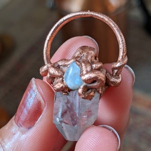 May include: A copper wire wrapped around a clear quartz crystal with a blue moonstone accent. The crystal is in the shape of a point and is held by a finger.