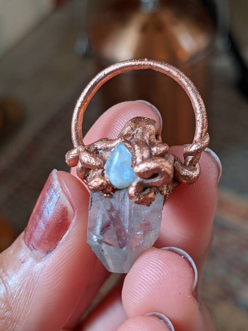 May include: A copper wire wrapped around a clear quartz crystal with a blue moonstone accent. The crystal is in the shape of a point and is held by a finger.