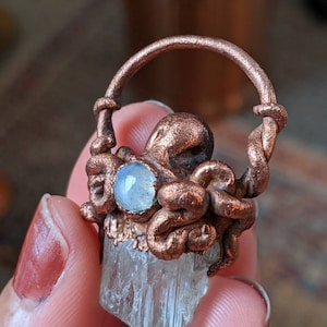 May include: A copper octopus charm with a clear crystal and a moonstone pendant. The charm is attached to a copper ring.