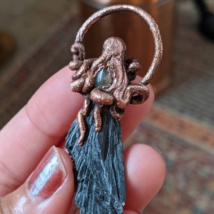 May include: A copper wire octopus pendant with a green gemstone in the center, hanging from a copper wire circle. The octopus is attached to a black, striated, crystal pendant.