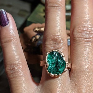 May include: A ring with a large, oval-shaped green gemstone set in a copper band. The gemstone has a rough, textured surface and is surrounded by a decorative copper border.