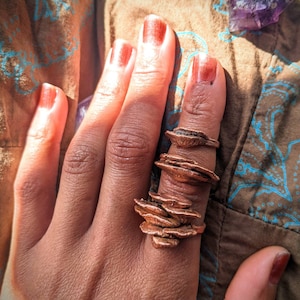 Puede incluir: Una mano con una pila de anillos de cobre con un diseño orgánico en capas. Los anillos están en el dedo índice y están hechos de varias piezas de metal apiladas una encima de la otra.