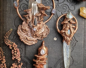 May include: Three copper and crystal pendants with a nature-inspired design. Each pendant features a clear quartz crystal and intricate copper details resembling branches and leaves. The pendants are displayed on a dark textured surface.