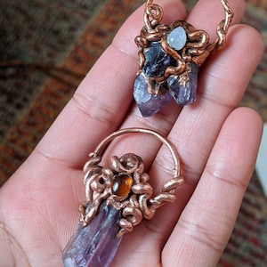 May include: Two amethyst crystal pendants wrapped in copper wire. One pendant features a small moonstone and the other features a small carnelian gemstone.