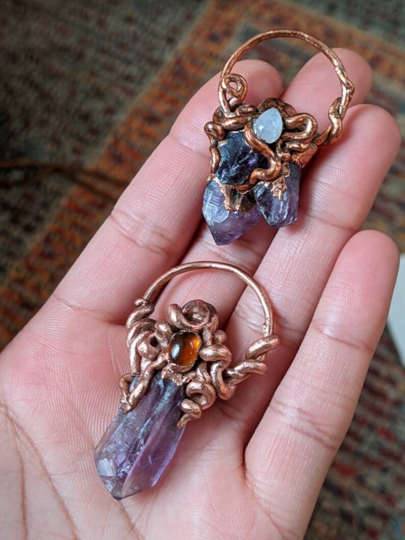 May include: Two amethyst crystal pendants wrapped in copper wire. One pendant features a small moonstone and the other features a small carnelian gemstone.
