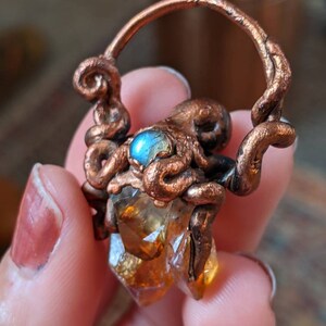 May include: A copper octopus pendant with a labradorite gemstone and a citrine crystal. The octopus is holding the crystal in its tentacles.