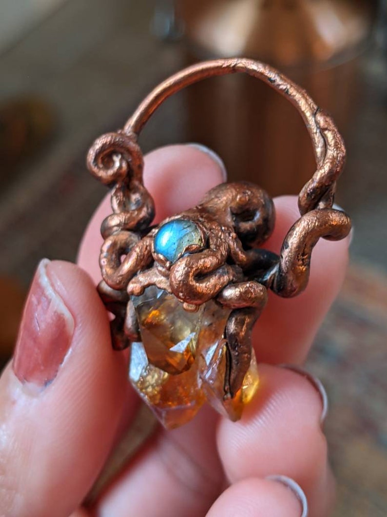 May include: A copper octopus pendant with a labradorite gemstone and a citrine crystal. The octopus is holding the crystal in its tentacles.