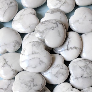 May include: A collection of white heart-shaped stones with gray veining. The stones are scattered on a light blue surface, creating a visually appealing arrangement. The heart-shaped stones are smooth and polished.