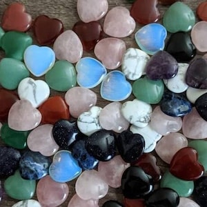 May include: A collection of heart-shaped polished stones in various colors, including pink, green, blue, purple, red, and black. The stones are arranged in a loose pile on a wooden surface.