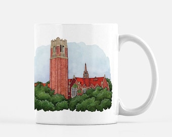 May include: White ceramic mug with a watercolor illustration of a red brick building with a tall tower and a bell tower. The building is surrounded by green trees.