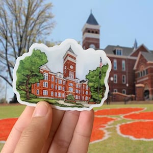 May include: A sticker of a red brick building with a clock tower and a flagpole. The building is surrounded by green trees and a grassy lawn. The sticker is held in a hand against a background of a red and white pattern.