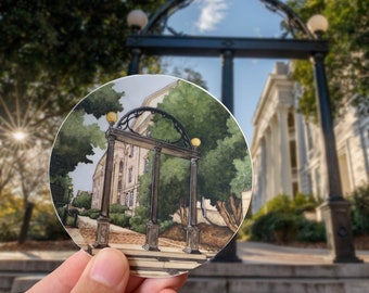 May include: A round sticker with a watercolor illustration of the iconic archway at the University of Georgia. The archway is painted in a dark brown color with a light brown brick pathway leading up to it. The archway is surrounded by green trees and a building in the background.