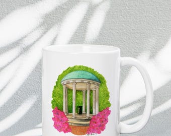 May include: White ceramic mug with a watercolor illustration of a white gazebo with a green roof surrounded by pink flowers. The gazebo is on a green lawn.