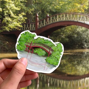 May include: A sticker featuring a watercolor illustration of a red bridge over water, surrounded by green trees. The bridge has a curved design with a white railing. The sticker is held in front of a blurred background of a similar bridge and trees.