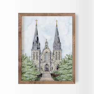 May include: Watercolor painting of a church with two tall spires topped with gold crosses. The church is made of stone and has a detailed facade. The artwork is framed in a rustic wooden frame, with green trees on either side.