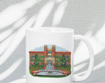 May include: White ceramic coffee mug with a colorful illustration of a brick building with a fountain in front. The building has a red roof and white windows. The fountain has a water spout and a pool of water. The text "Florida State University" is printed below the fountain.