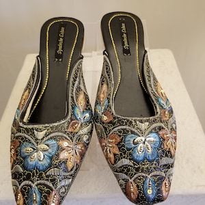 Może przedstawiać: Para czarnych, haftowanych kapci z nitką złotą i srebrną. Kapcie mają spiczasty nosek i niski obcas. Hafty przedstawiają kwiatowy wz&oacute;r z niebieskimi, złotymi i brązowymi akcentami. Nazwa marki "Syndeeche Esham" jest nadrukowana na wkładce.