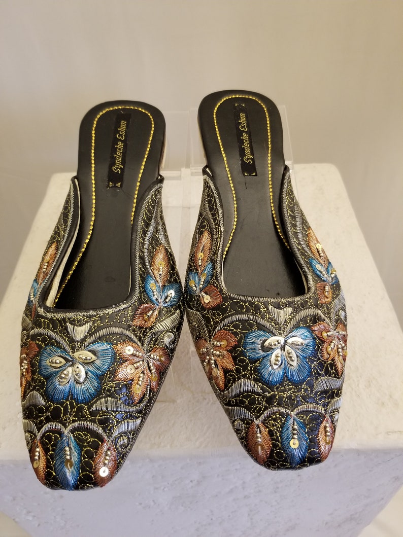 Może przedstawiać: Para czarnych, haftowanych kapci z nitką złotą i srebrną. Kapcie mają spiczasty nosek i niski obcas. Hafty przedstawiają kwiatowy wz&oacute;r z niebieskimi, złotymi i brązowymi akcentami. Nazwa marki "Syndeeche Esham" jest nadrukowana na wkładce.