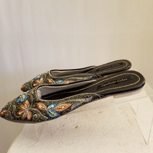 Może przedstawiać: Para czarnych, haftowanych kapci z złotymi i niebieskimi akcentami. Kapcie mają spiczasty nosek i płaską podeszwę. Nazwa marki "Syndeeche Eshun" jest nadrukowana na podeszwie.