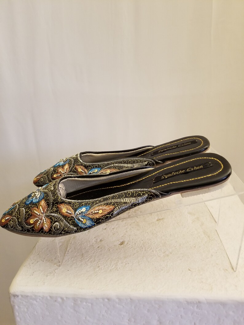 Może przedstawiać: Para czarnych, haftowanych kapci z złotymi i niebieskimi akcentami. Kapcie mają spiczasty nosek i płaską podeszwę. Nazwa marki "Syndeeche Eshun" jest nadrukowana na podeszwie.